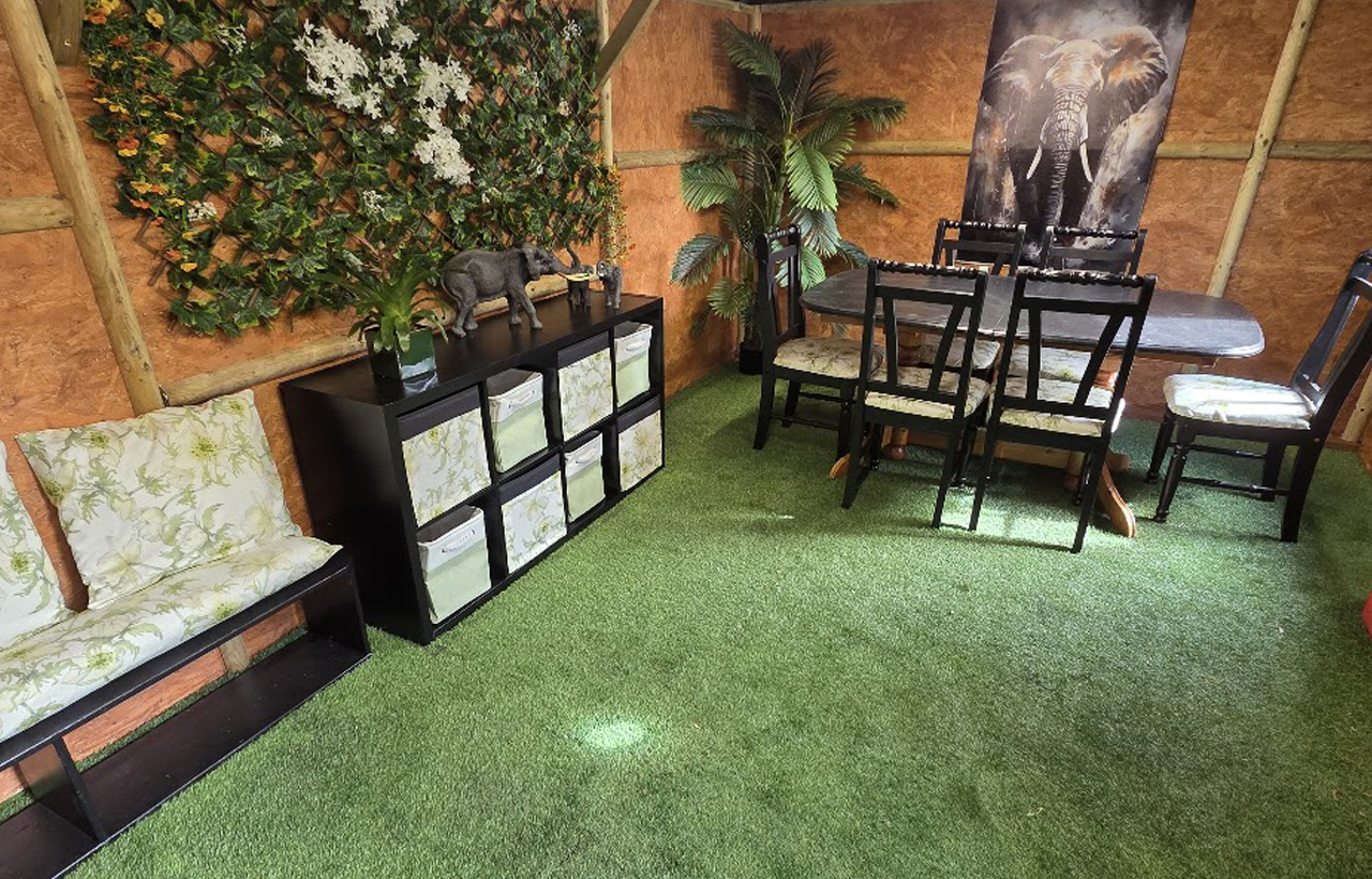 A room with artificial grass flooring, a table with six chairs, a black storage unit, wall art of an elephant, decorative plants, and a cushioned bench.