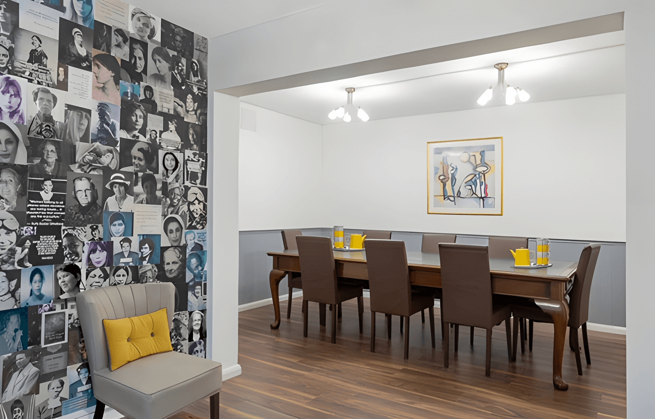 A modern conference room with a wooden table, brown chairs, and abstract art on the wall; a collage of black-and-white portraits covers one wall.