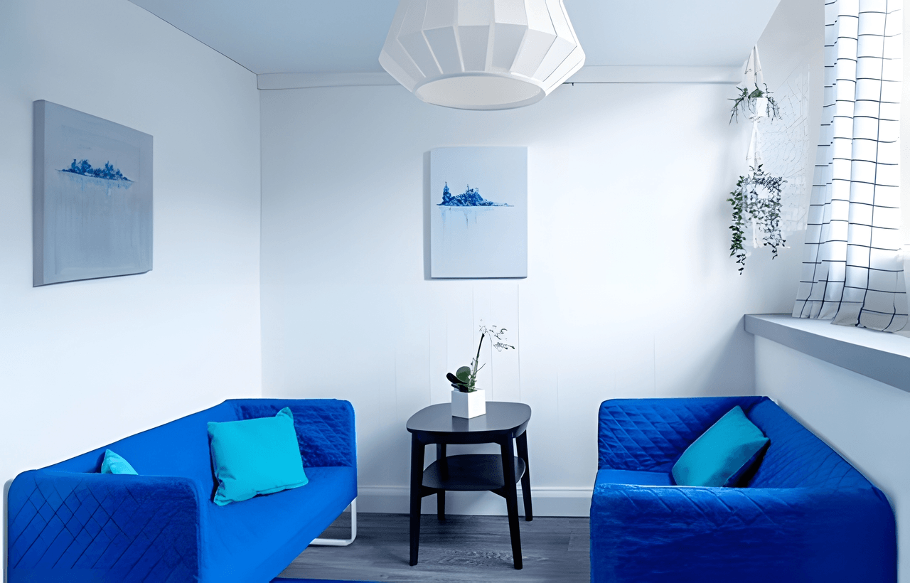 A small, modern sitting area with two blue sofas, a black table with a white potted plant, wall art, a hanging lamp, and a window with sheer curtains.