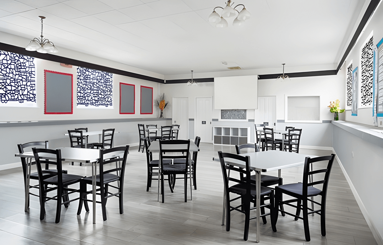 A clean, modern hall with black chairs and white tables, wall art, large windows, and light fixtures on the ceiling.