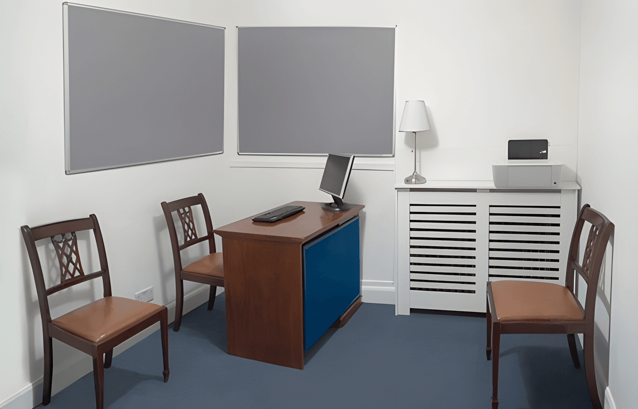 A small office with a desk, computer, keyboard, three chairs, two bulletin boards, a lamp, and a printer on a side cabinet.